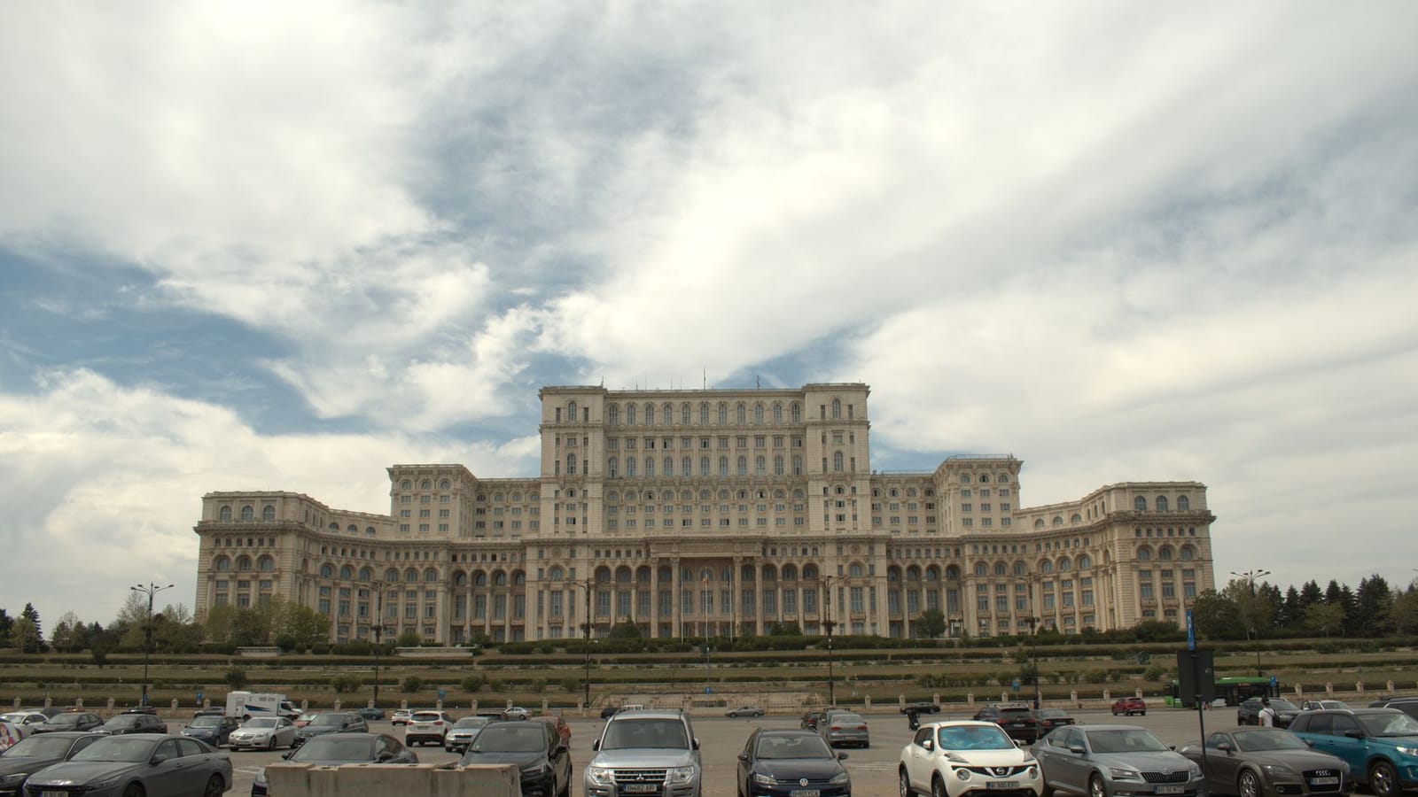 A daytime view of the iconic Palace of the Parliament in Bucharest, Romania, featuring the grand architecture and parked cars.