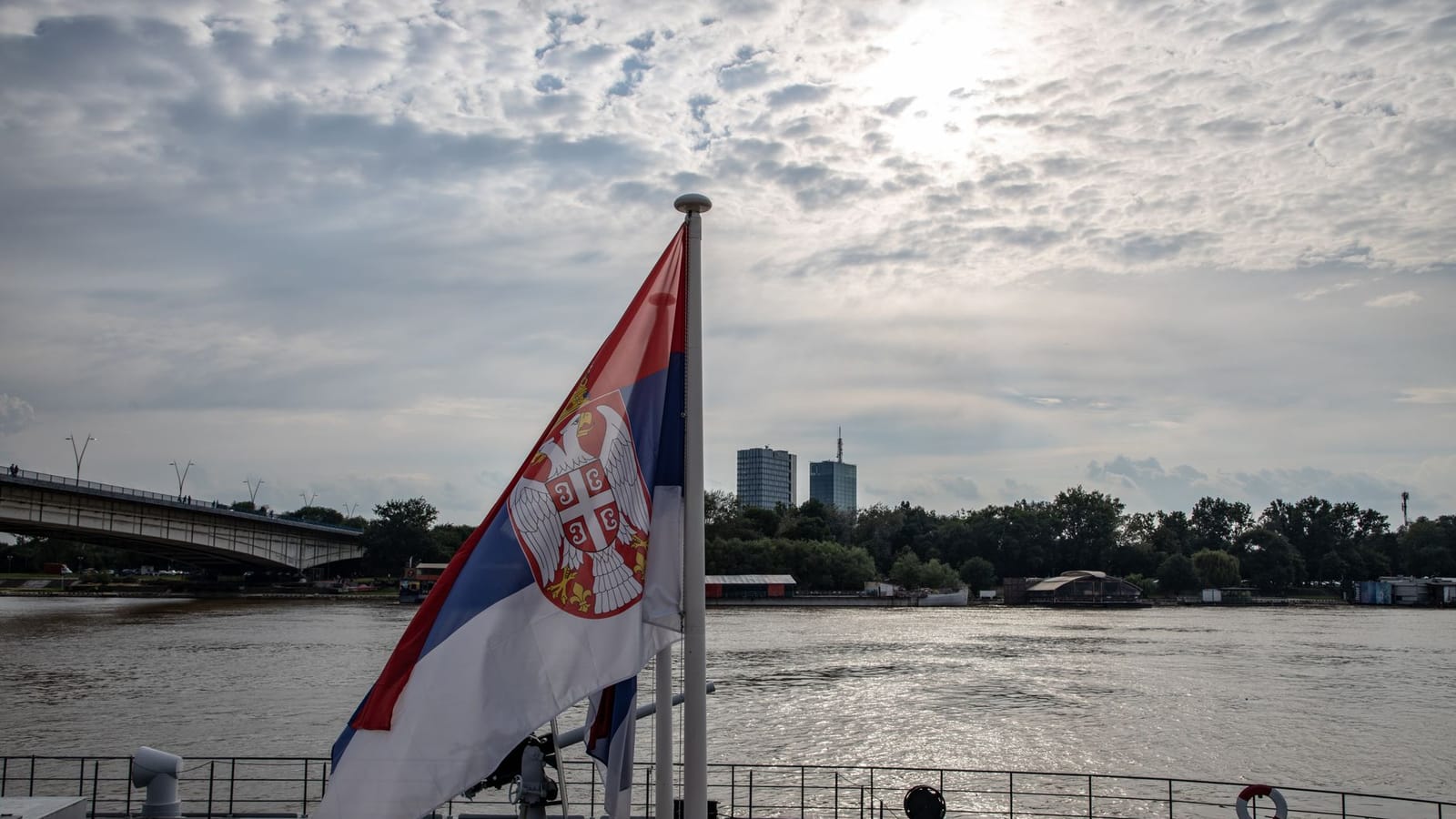 Serbian flag on a boat with an urban skyline and river scene in daytime.