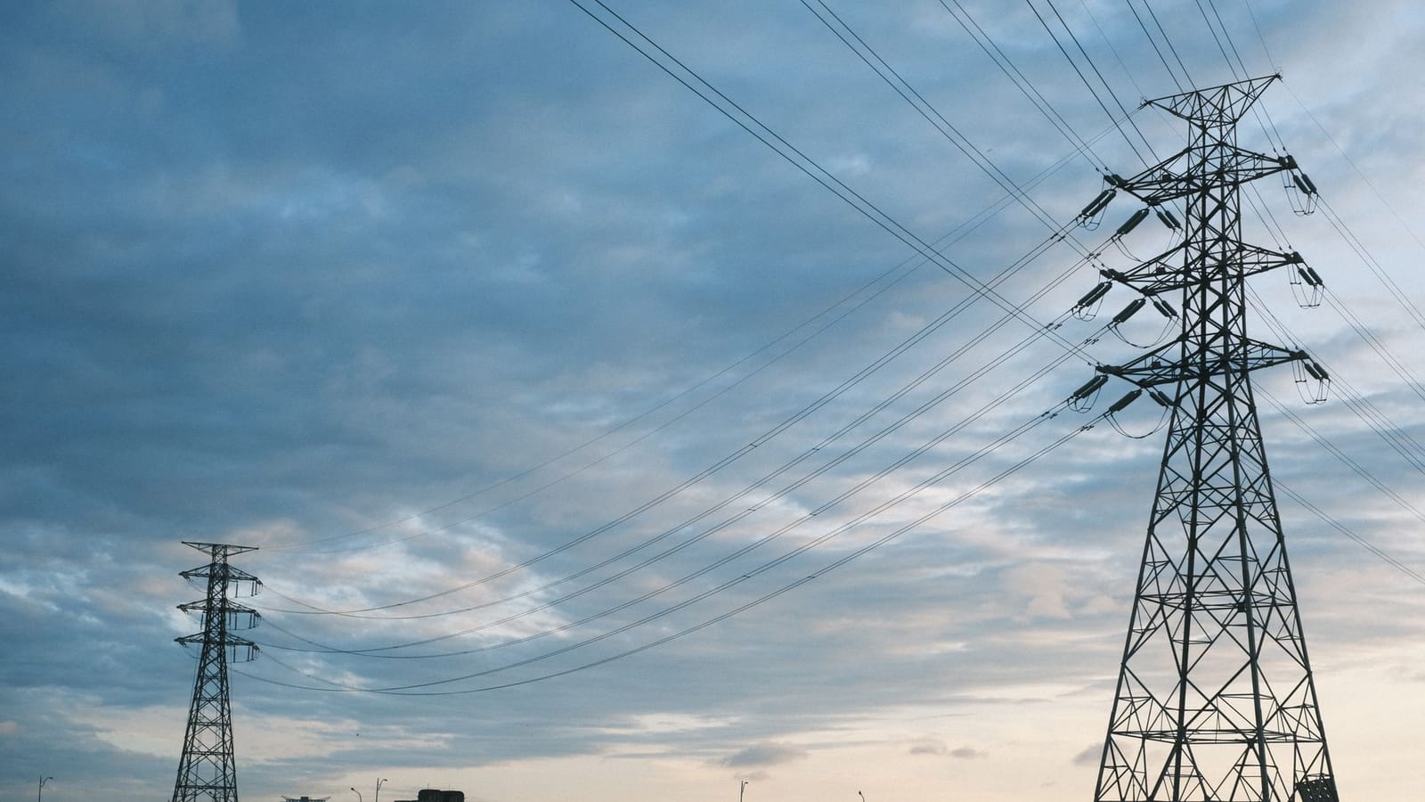A city skyline at dusk with transmission towers and power lines silhouetted against a cloudy sky.