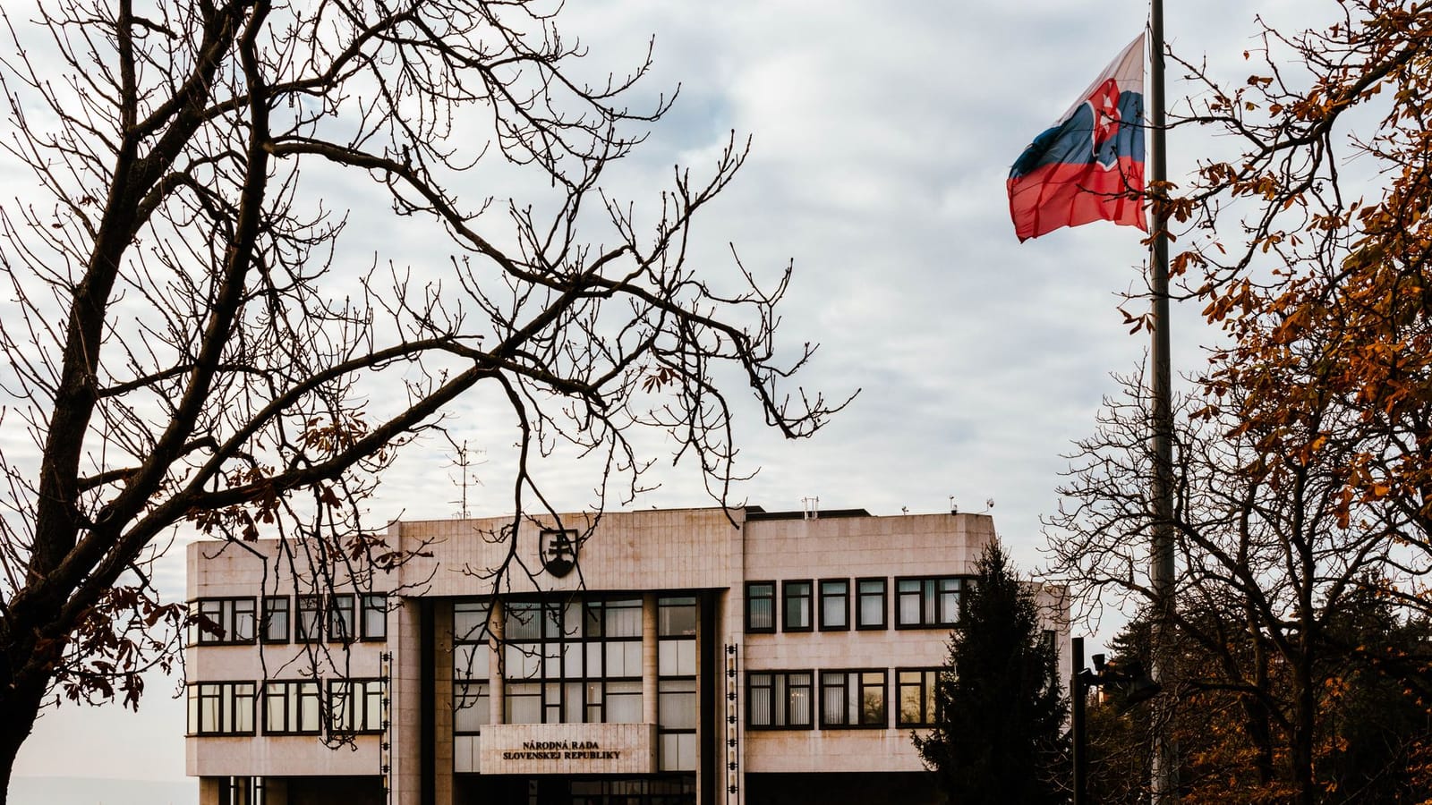A view of the Slovak Parliament building surrounded by autumn trees with the national flag waving under an overcast sky.
