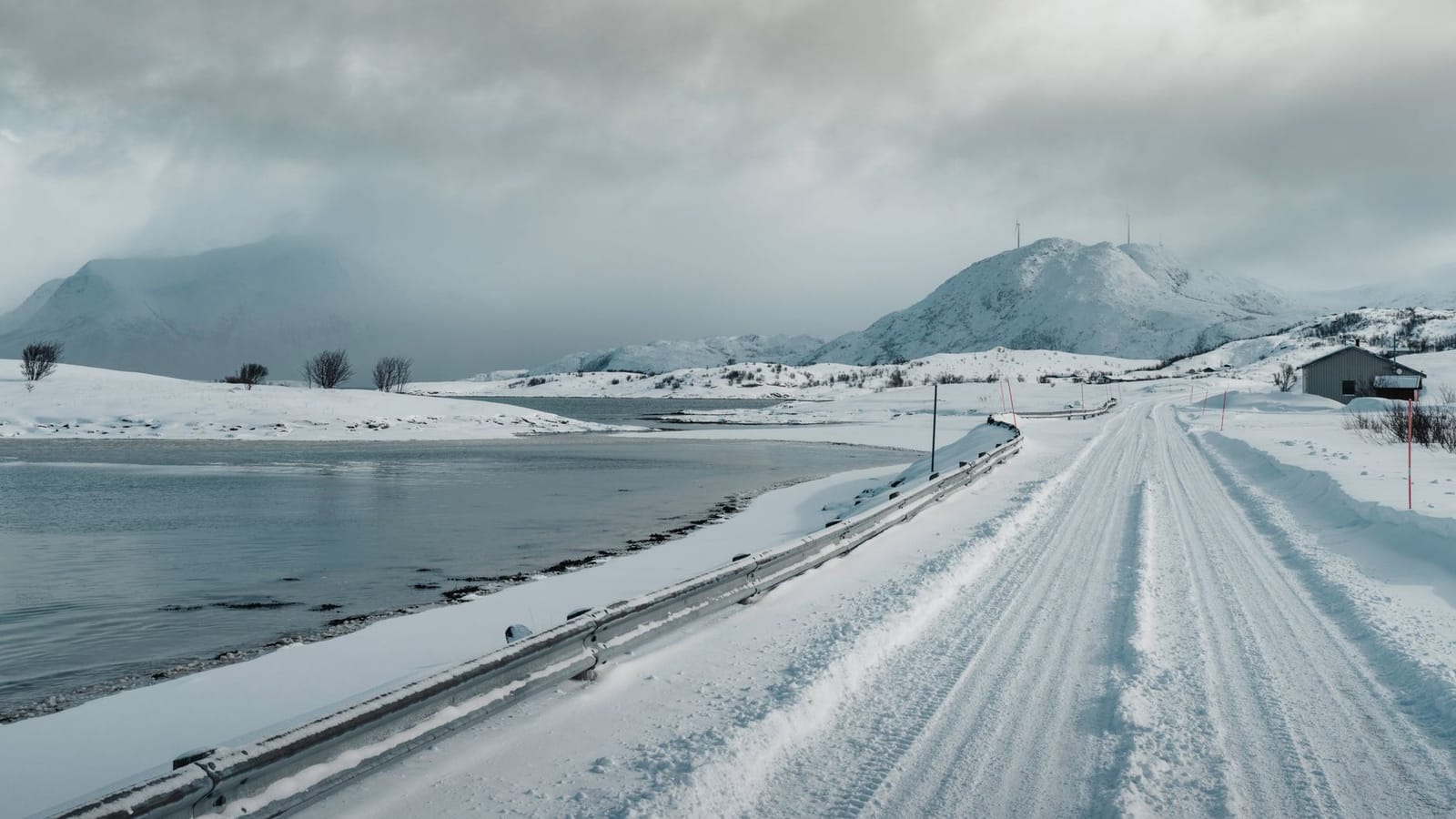 Snow-covered road in Norway with mountains and a lake under cloudy skies.