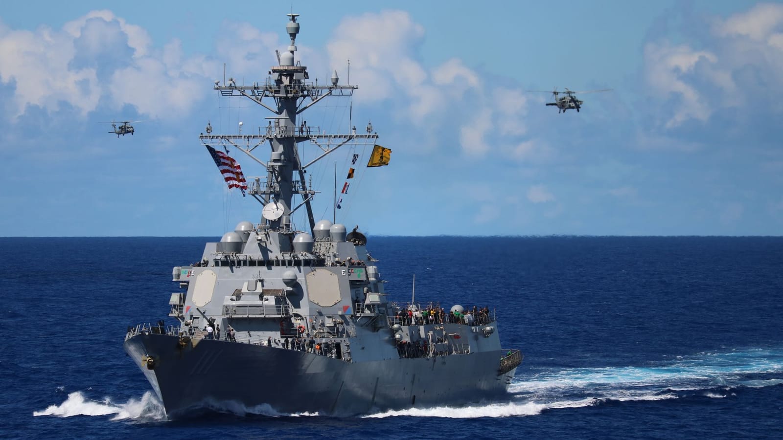 A US Navy destroyer navigates the ocean with helicopters in formation above.