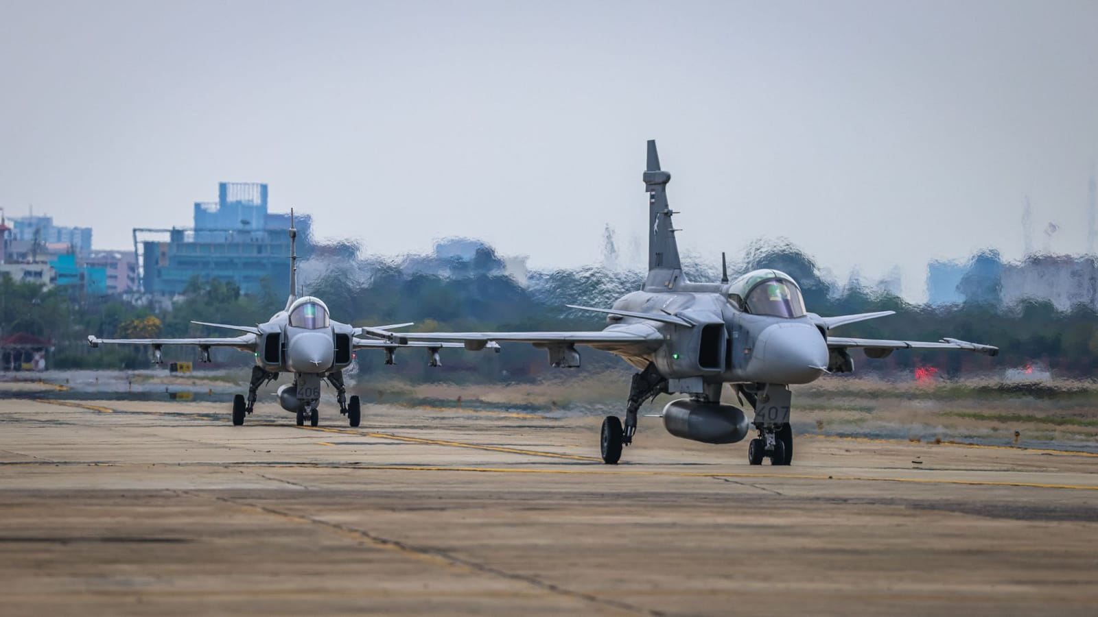 Two military fighter jets taxiing on the runway, cityscape in the background.