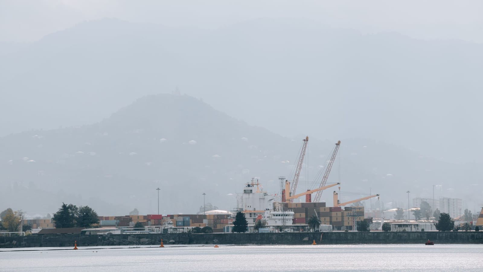 A foggy port scene with cranes, shipping containers, and mountains in the background.