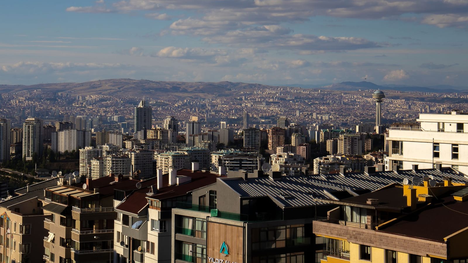 A sweeping aerial view of Ankara's cityscape under a bright, cloudy sky.