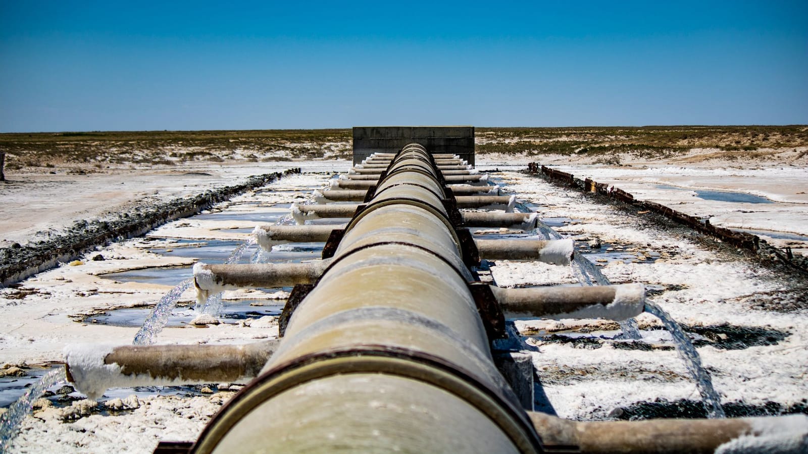 Large industrial pipeline discharging wastewater in arid, rural landscape under clear blue sky.
