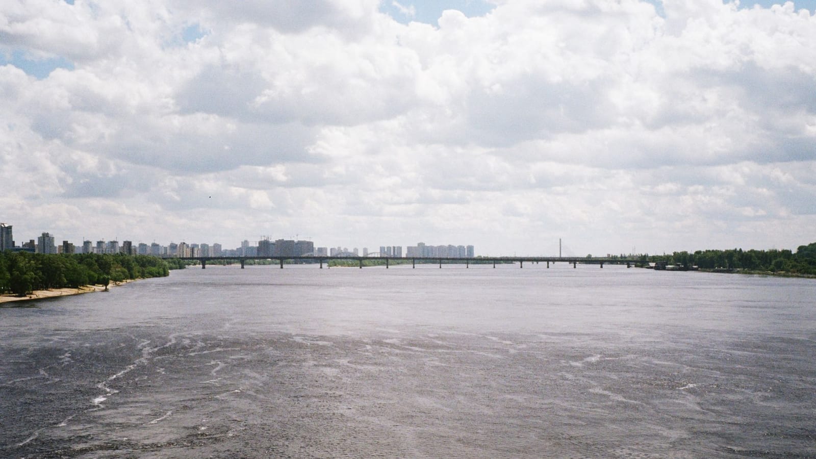 A panoramic view of the Dnipro River with a bridge and Kyiv city skyline under a cloudy sky.