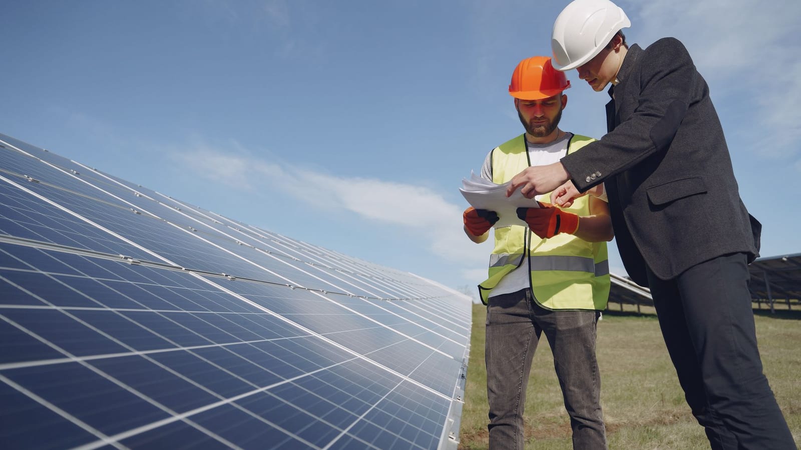 Low angle of young inspector and foreman in hardhats checking documentation against modern solar panels in field