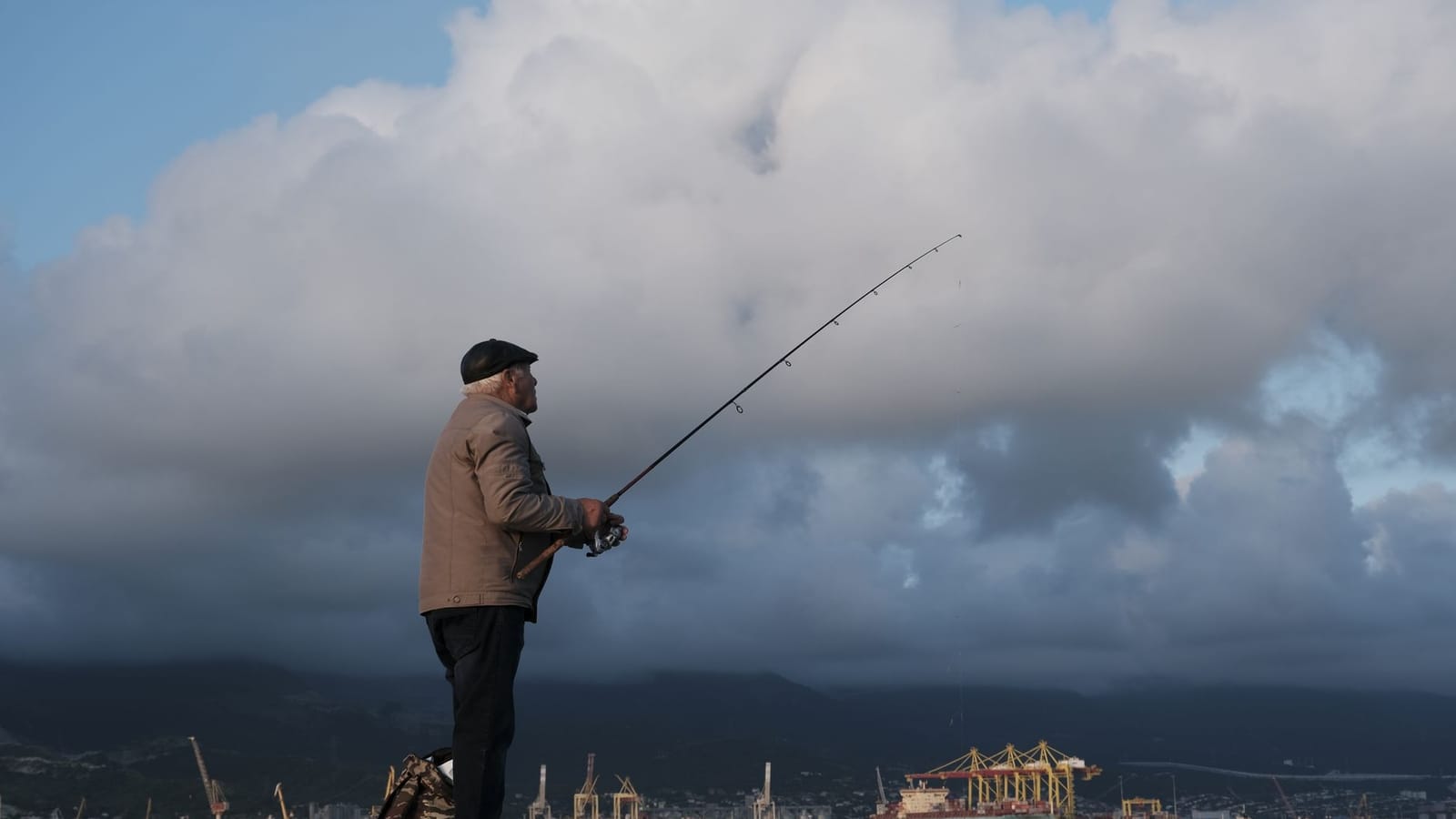 An elderly man fishing by Novorossiysk port amidst dramatic clouds.