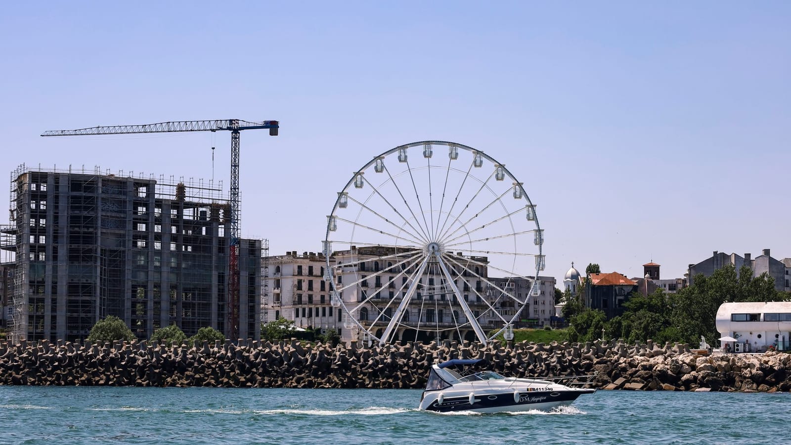 A sunny day view in Constanța with a ferris wheel, construction site, and a boat in the foreground.