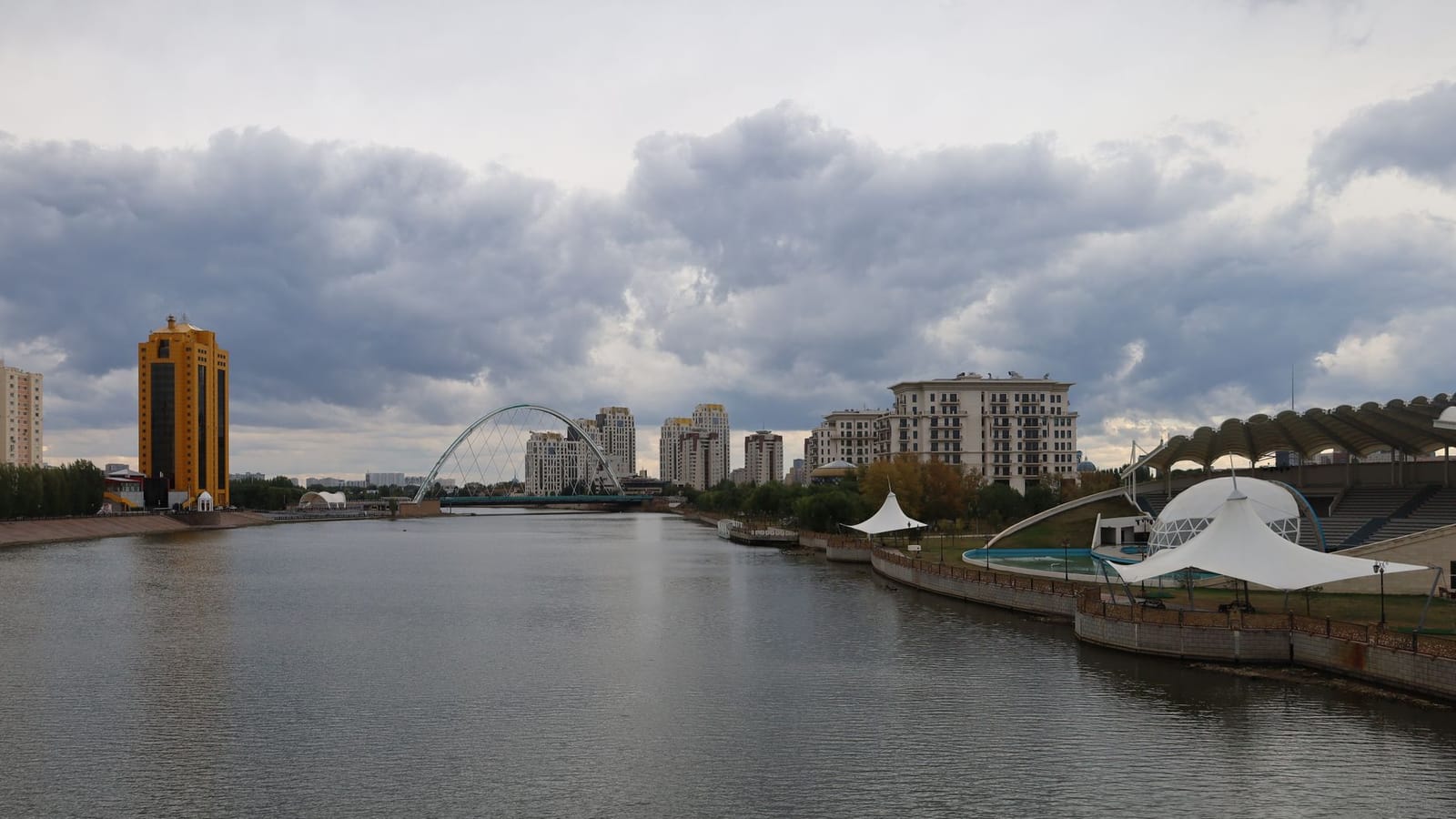 Modern skyline of Astana with residential buildings along the Ishim River under cloudy skies.