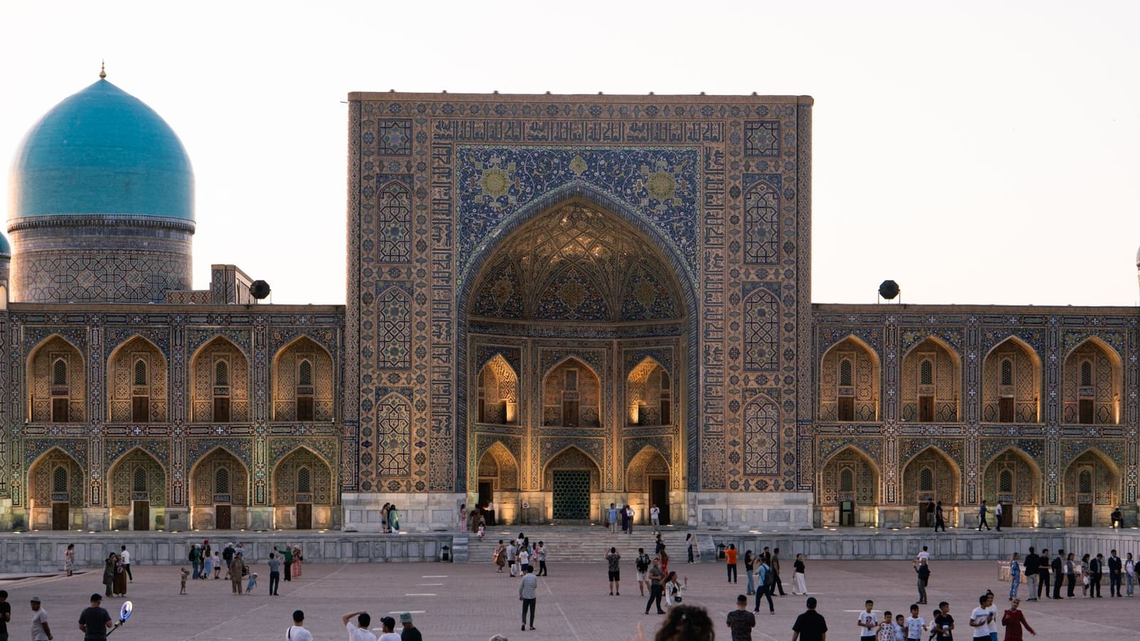 People gathering at the illuminated Registan complex in Samarkand at dusk.