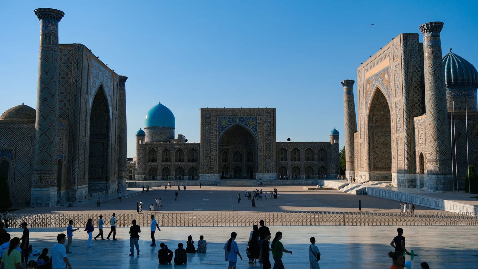 Crowded Registan Square in Samarkand with ancient madrasahs under a clear sky.