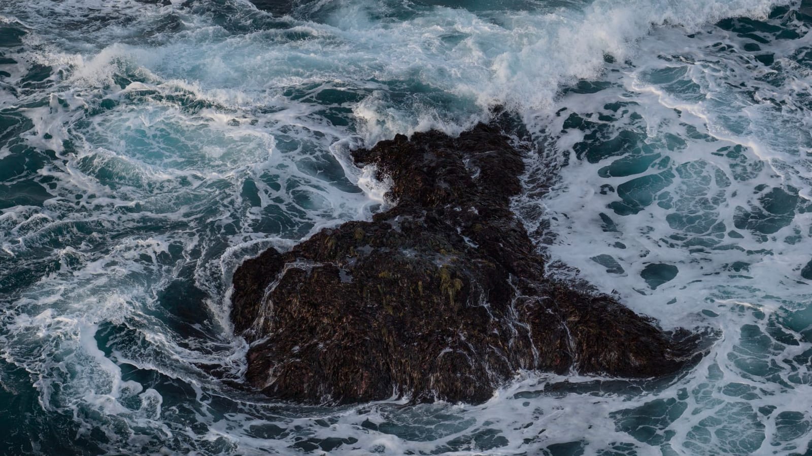 A dynamic aerial photo capturing ocean waves crashing on a rugged sea rock.