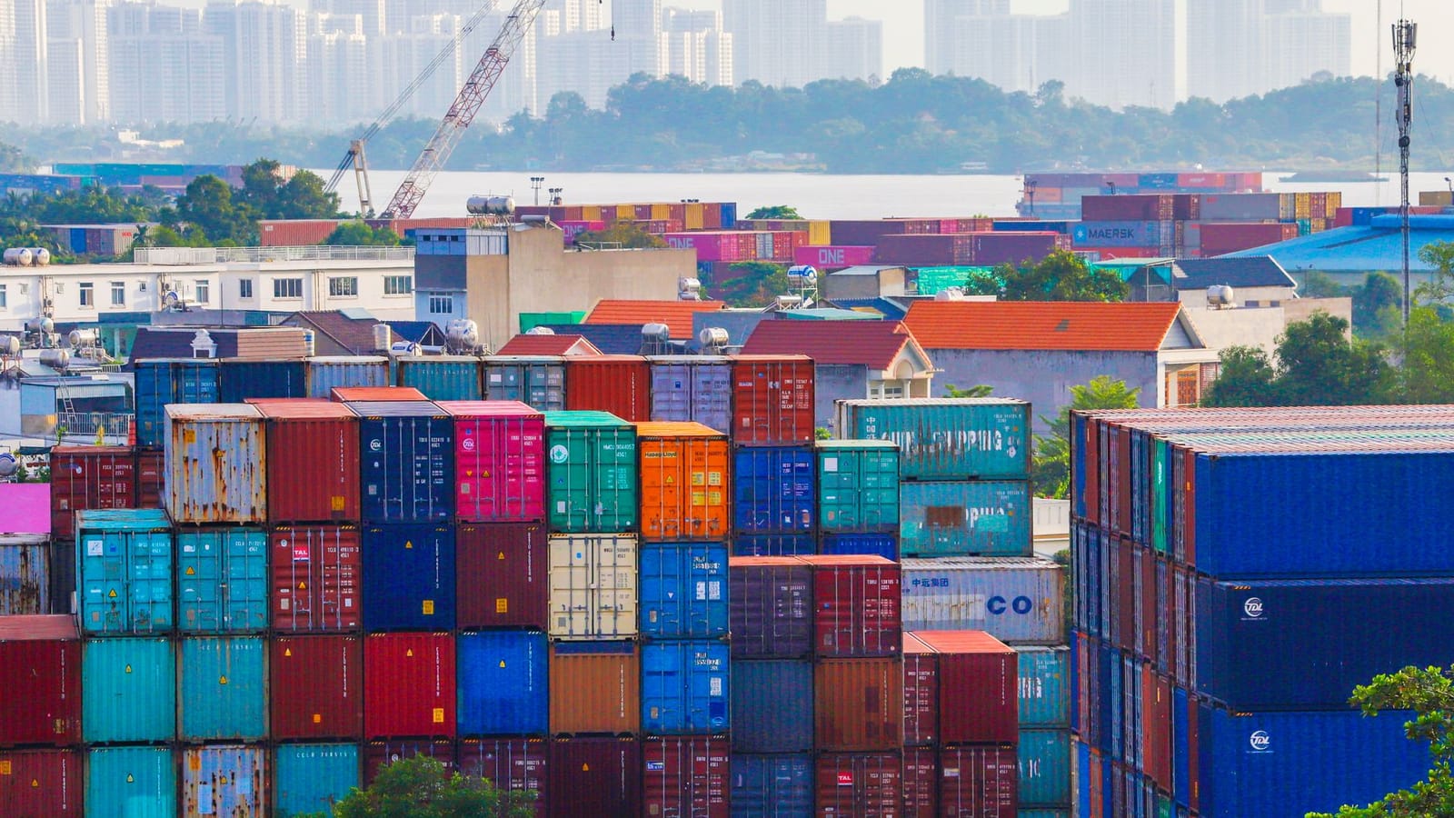 Vibrant stacked cargo containers at a bustling port with urban skyline in background.