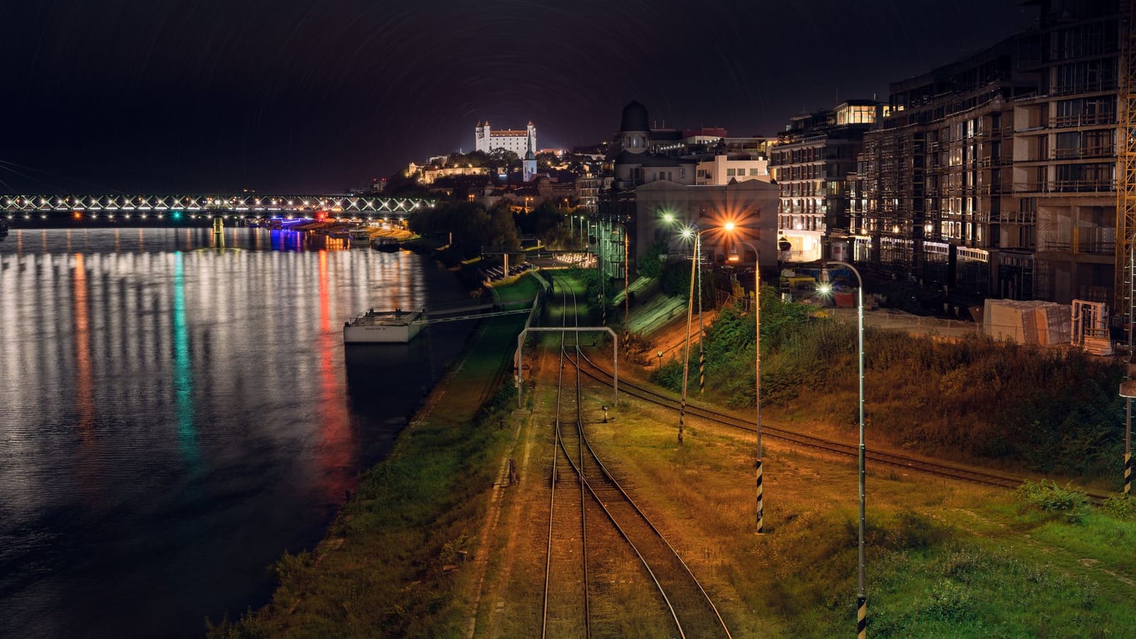 Scenic night shot of Bratislava with river reflections, railway tracks, and the illuminated castle.