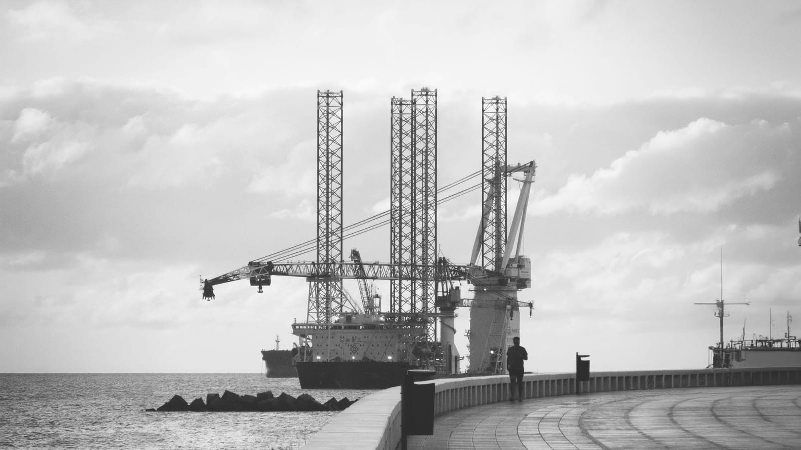 Monochrome shot of a coastal oil rig near a curved walkway under cloudy skies.