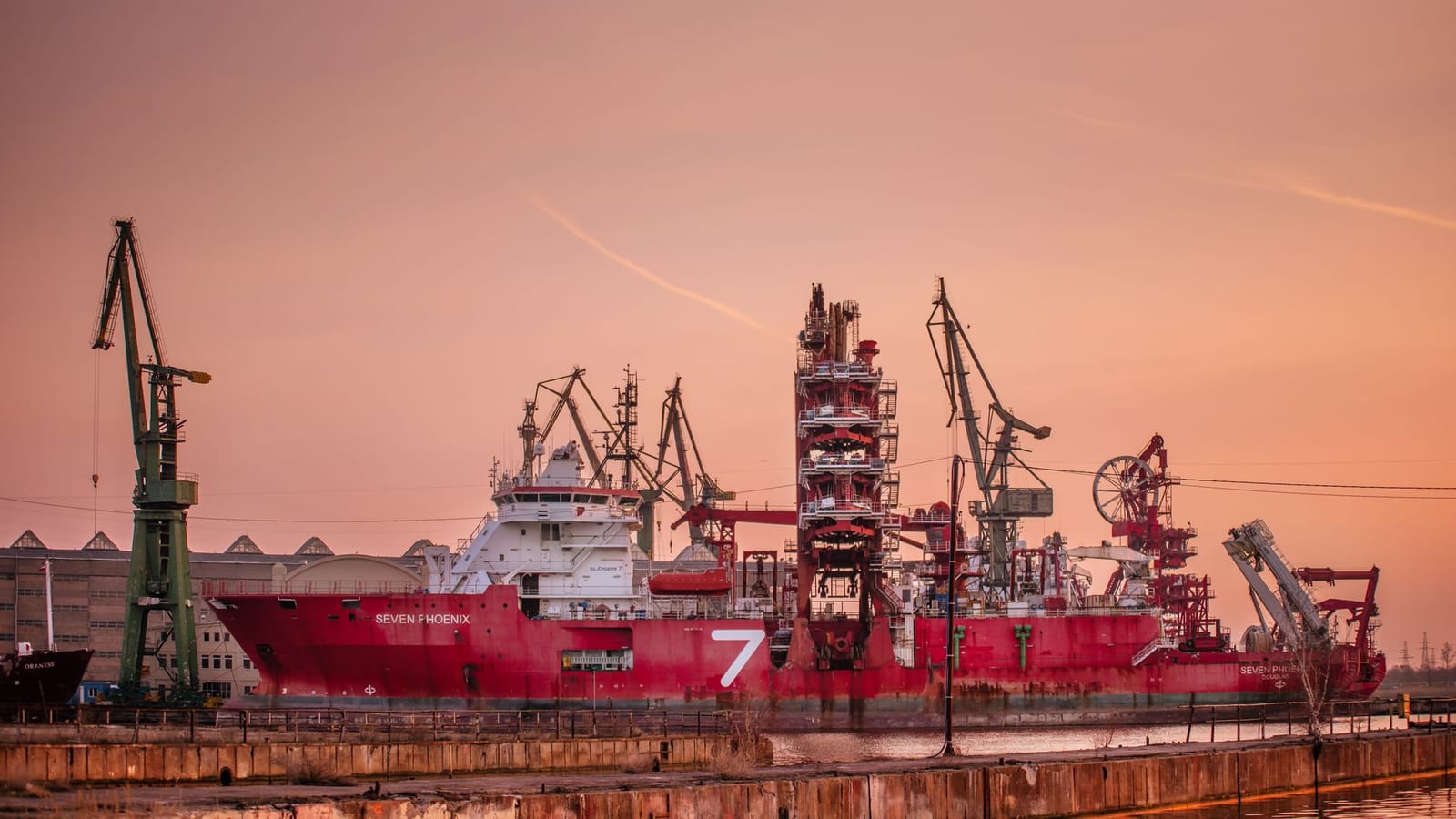 Industrial ship moored in Gdańsk harbor at sunset with cranes in view.