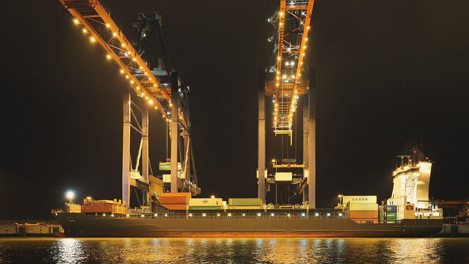 Container ship docked in Rotterdam Harbor illuminated by cranes at night, showcasing industrial infrastructure.