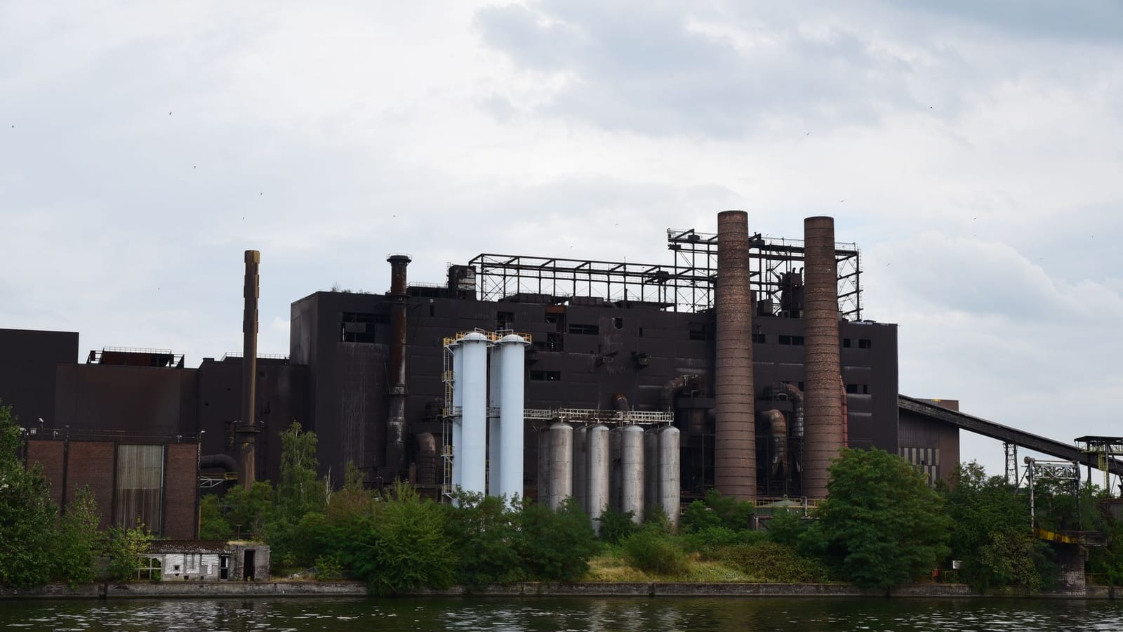 A large industrial factory with chimneys by a river, surrounded by trees on a cloudy day.