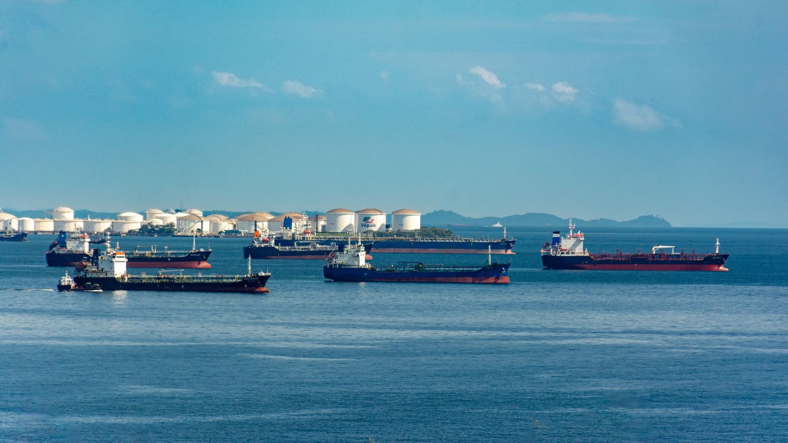 A fleet of cargo ships docked near oil storage tanks along a serene coastline with a clear blue sky above.
