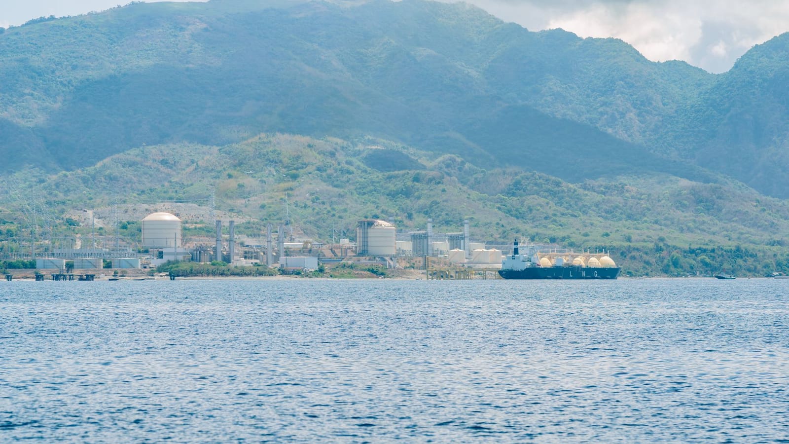 Industrial facility by the coast with a docked LNG tanker, set against mountain backdrop.