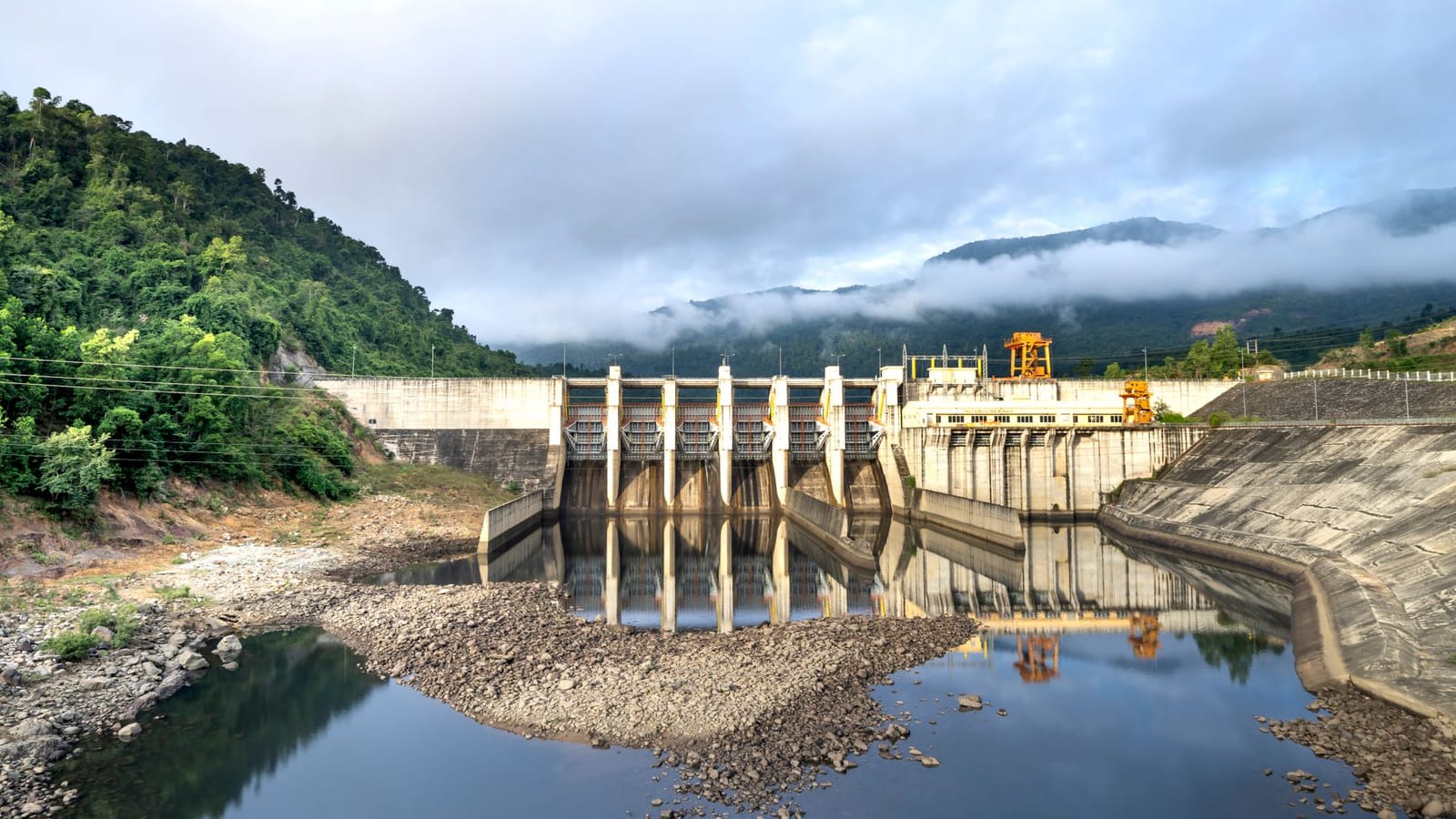 Industrial hydroelectric power plant located near river and stony coast against green mountain range covered with mist on summer day