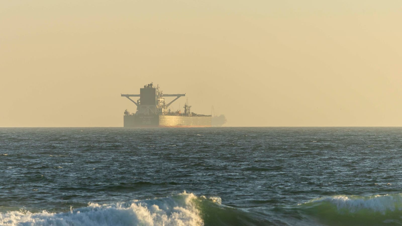 A cargo ship silhouetted against the horizon on a calm ocean during twilight.