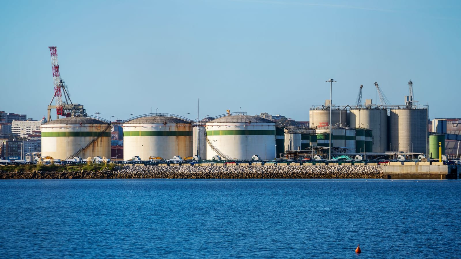 Large storage tanks at an industrial port facility by the water under clear blue skies.